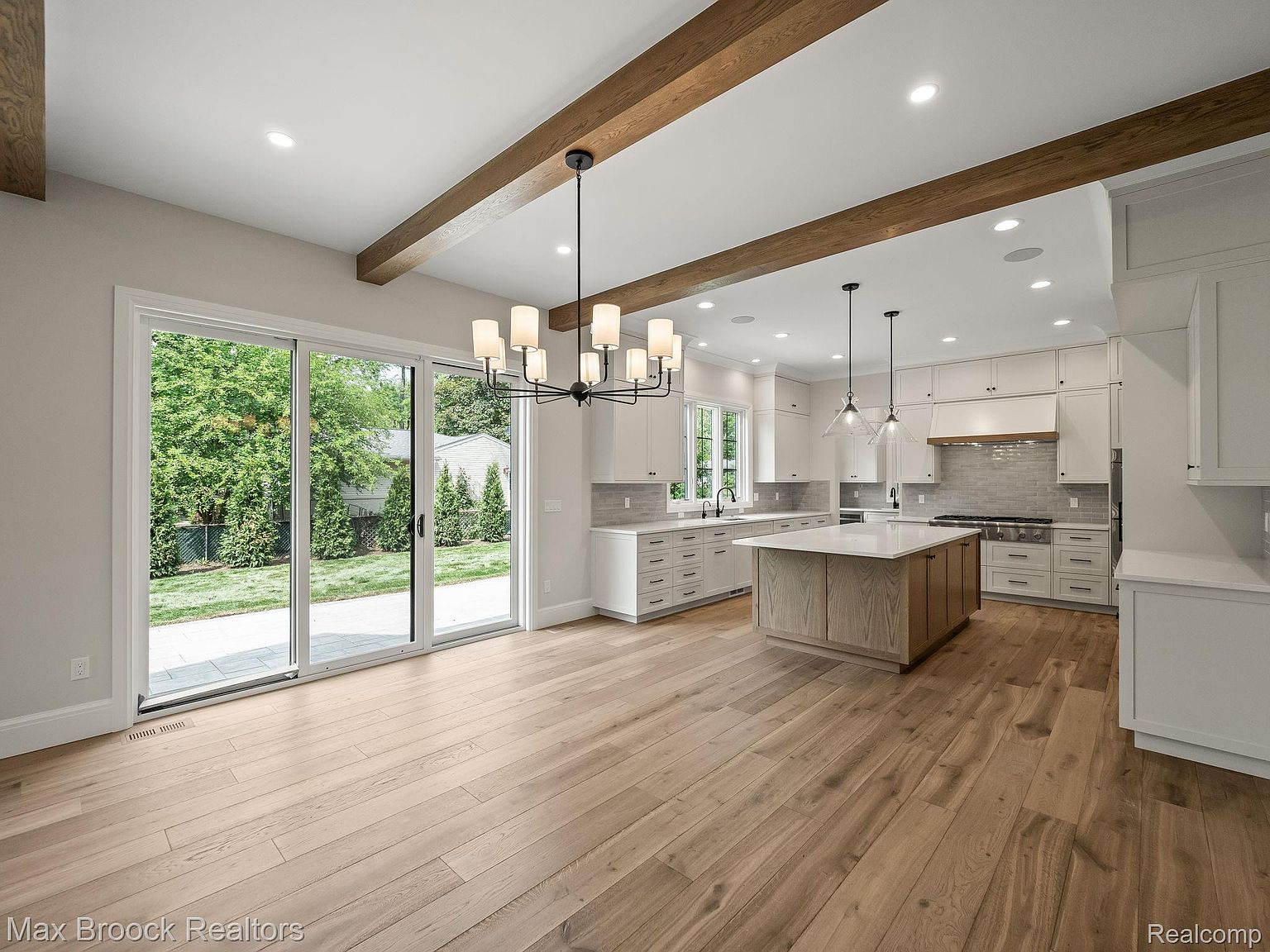 This is a bright and spacious kitchen featuring a large center island with a light wood base and white countertop. The kitchen is equipped with white cabinetry, stainless steel appliances, and modern pendant lighting. Large sliding glass doors provide ample natural light and a view of the backyard, enhancing the open and airy feel of the space.