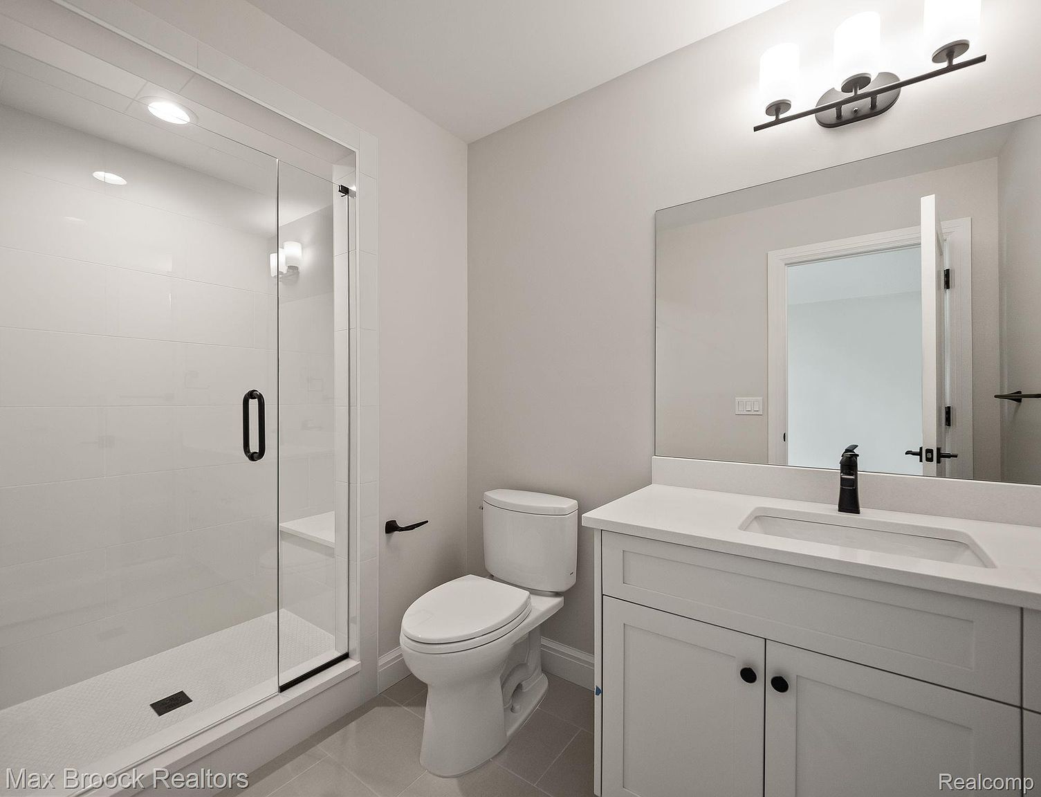 This is a well-lit bathroom featuring a glass-enclosed shower with white tile, a toilet, and a vanity with a white countertop and dark hardware. The walls are painted in a neutral tone, and the overall design is clean and modern. A large mirror reflects the room, enhancing the sense of space.