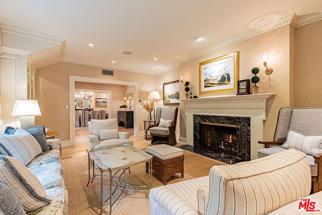 This is a warm and inviting living room featuring a fireplace with a marble surround and a white mantel. The room is furnished with comfortable seating, including sofas and armchairs, arranged around a coffee table. The walls are adorned with artwork, and the overall style is traditional and elegant, creating a cozy and sophisticated atmosphere.