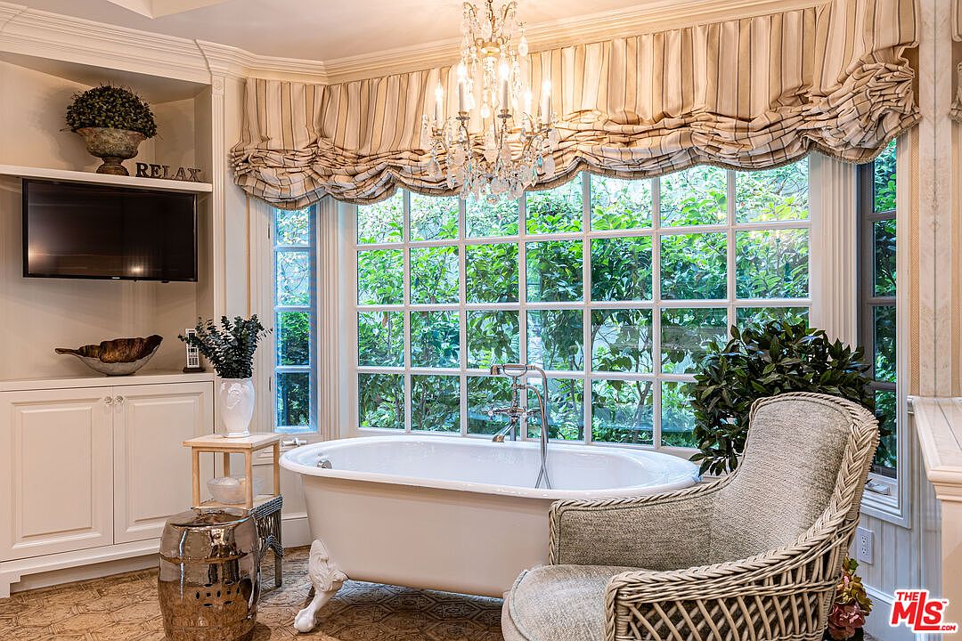 This is a luxurious primary bathroom featuring a clawfoot bathtub positioned in front of a large window offering a view of lush greenery. The room is adorned with a crystal chandelier, custom window treatments, and a comfortable armchair, creating a spa-like atmosphere. Built-in cabinetry and a wall-mounted television add to the room's functionality and appeal.