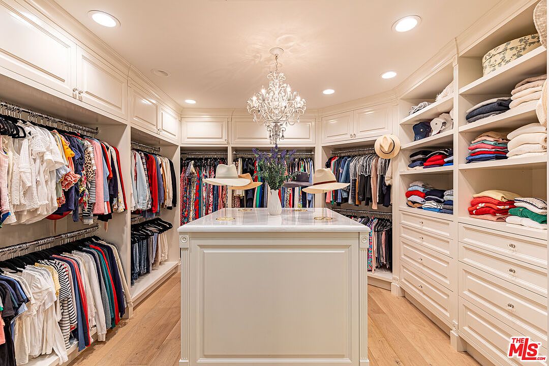 This is a well-organized walk-in closet featuring custom white cabinetry and shelving. Clothes are neatly hung and folded, and a central island provides additional storage and display space for hats. A chandelier adds a touch of elegance to the space, which is illuminated by recessed lighting and features light hardwood flooring.