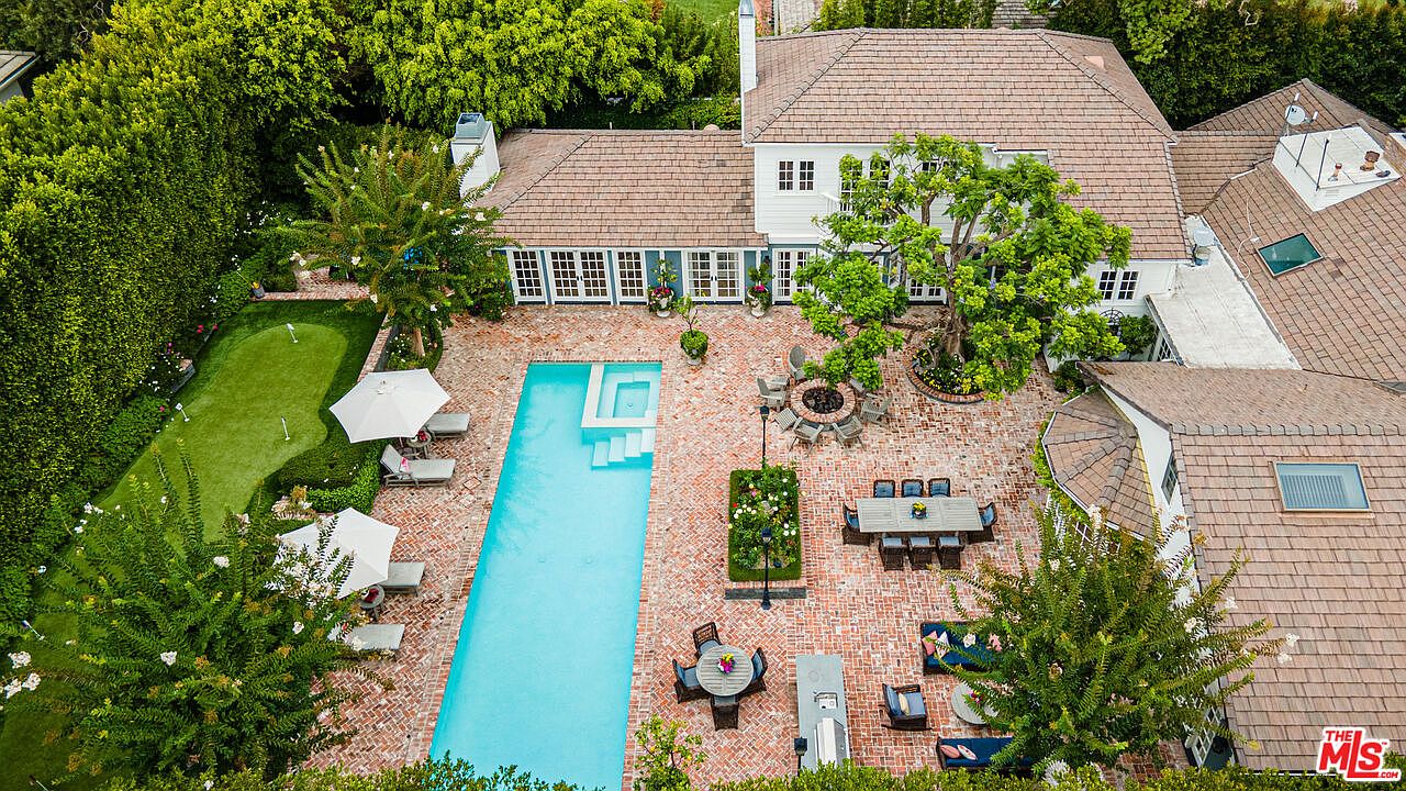 This aerial view showcases a luxurious backyard featuring a long rectangular pool with a built-in spa, surrounded by a brick patio. The outdoor space includes a putting green, multiple seating areas with tables and lounge furniture, and a fire pit, all set against a backdrop of lush greenery and a well-maintained white house with a brown tile roof. The overall impression is one of sophisticated outdoor living and relaxation.