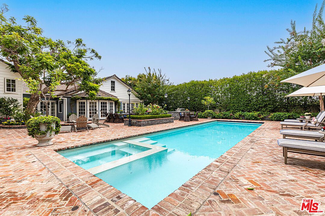 This image showcases a luxurious backyard pool and spa area. The pool is surrounded by a brick patio, with lounge chairs and an umbrella providing a relaxing space. The property features lush landscaping and a charming house in the background, creating a serene and private oasis.