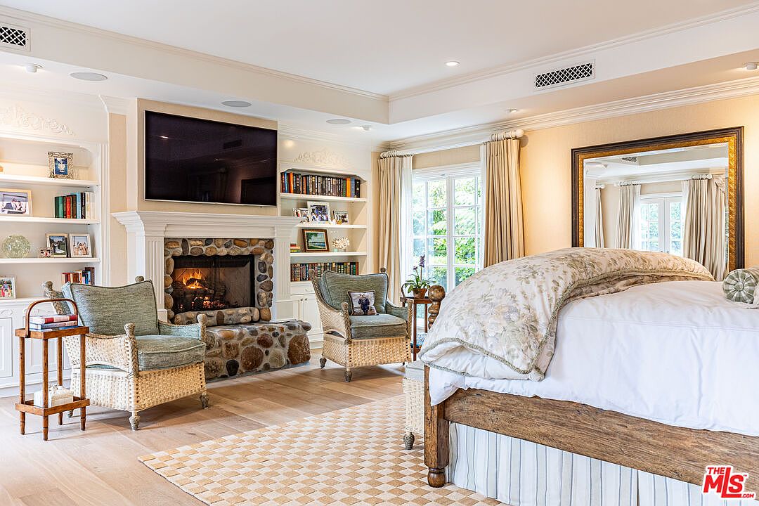 This is a primary bedroom featuring a stone fireplace with a mounted television above, flanked by built-in bookshelves. Two wicker chairs with cushions face the fireplace, and a large bed with a patterned duvet is positioned in the foreground. The room has hardwood floors and a neutral color palette, creating a cozy and inviting atmosphere.