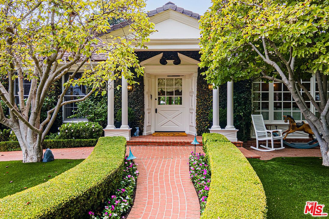 This is a charming front entryway of a home, featuring a brick pathway leading to a white front door framed by white columns and lush greenery. The manicured hedges and flowering plants add to the curb appeal, creating a welcoming and picturesque scene. A rocking chair and rocking horse can be seen to the right of the door.