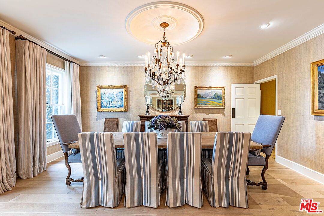 This is an interior shot of a dining room featuring a long wooden table surrounded by upholstered chairs with striped slipcovers. A chandelier hangs above the table, and artwork adorns the walls, creating a sophisticated and inviting atmosphere. The room is well-lit, highlighting the hardwood floors and textured wallpaper.