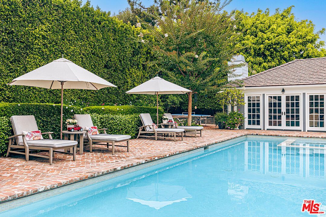 This image showcases a luxurious pool area, featuring a pristine blue pool surrounded by a brick patio. Lounge chairs with umbrellas offer a relaxing space, while lush greenery provides privacy and a serene backdrop. A building with multiple French doors is adjacent to the pool, suggesting easy indoor-outdoor living.