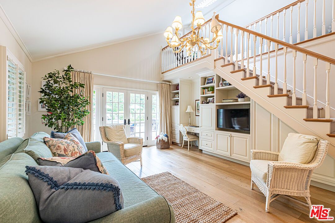 This is an interior shot of a living room featuring a light blue sofa with decorative pillows, two wicker chairs, and a jute rug on hardwood floors. The room has high ceilings, a staircase with white railings, and built-in shelving with a television. French doors lead to an outdoor area, and a chandelier hangs from the ceiling, creating a bright and inviting atmosphere.