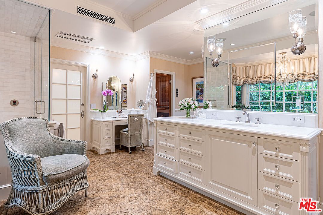 This is a well-lit primary bathroom featuring a large vanity with white cabinetry and a marble countertop. A decorative mirror and sconces adorn the wall above the vanity. The room includes a shower with glass doors, a comfortable armchair, and a separate vanity area with a chair, creating a luxurious and functional space.
