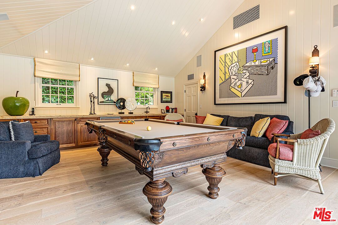 This interior shot showcases a well-appointed living room featuring a classic pool table as a central element. The room is decorated with a mix of modern and traditional elements, including a large pop art piece, comfortable seating, and custom cabinetry. Natural light filters through the windows, creating a warm and inviting atmosphere.