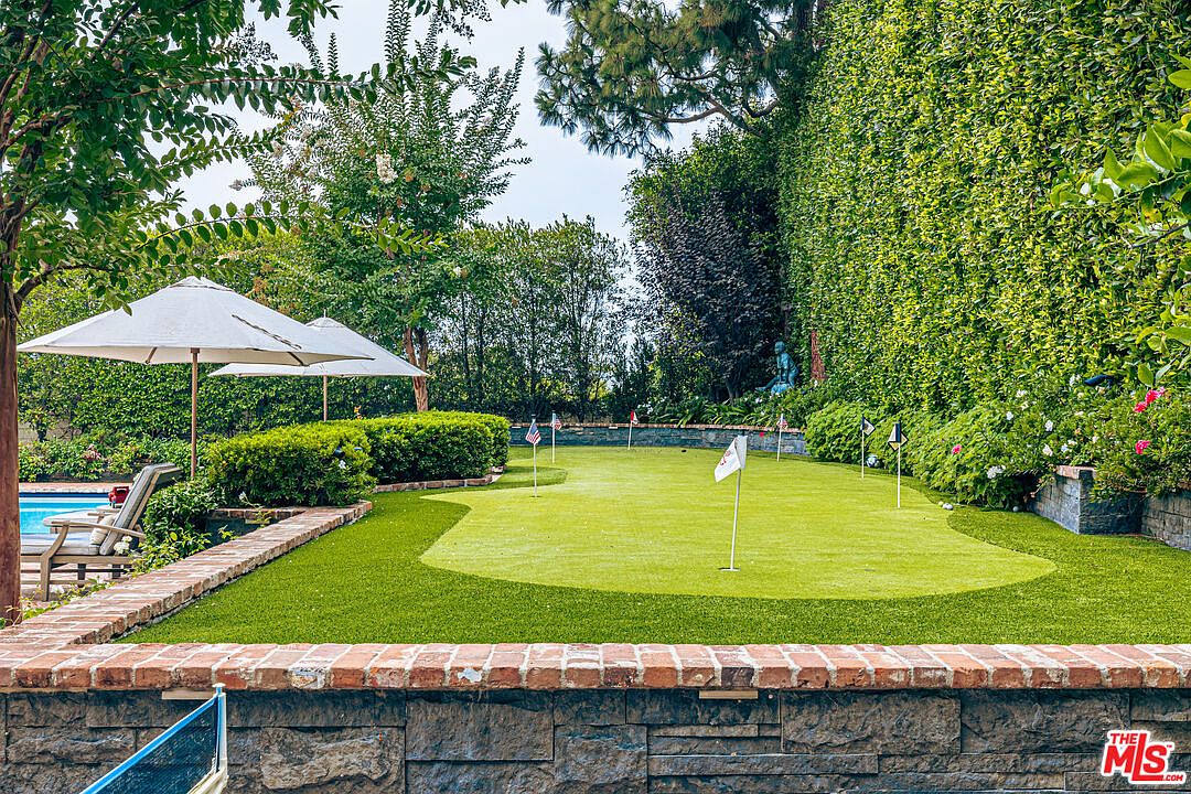 This image showcases a beautifully landscaped backyard featuring a putting green, lush greenery, and mature trees. A brick retaining wall adds texture and definition to the space, while patio umbrellas offer shade near the pool area. The overall impression is one of a private and well-maintained outdoor oasis.