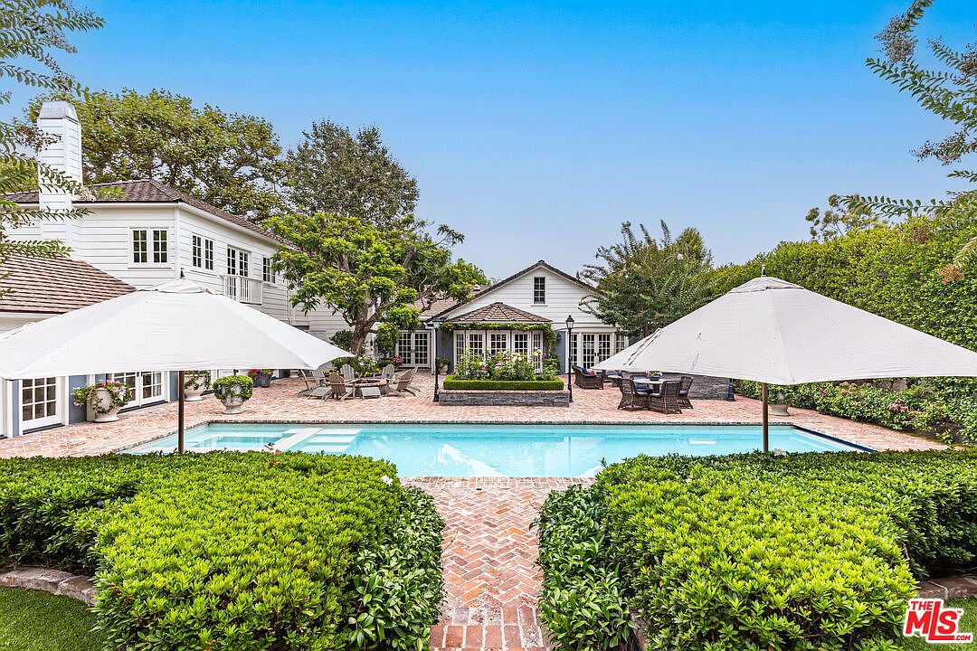 This image showcases a luxurious backyard featuring a pristine pool as the focal point. The pool is surrounded by a brick patio, lush greenery, and two large white umbrellas providing shade. The architecture of the house in the background suggests a classic, elegant style, enhancing the overall appeal of the outdoor living space.