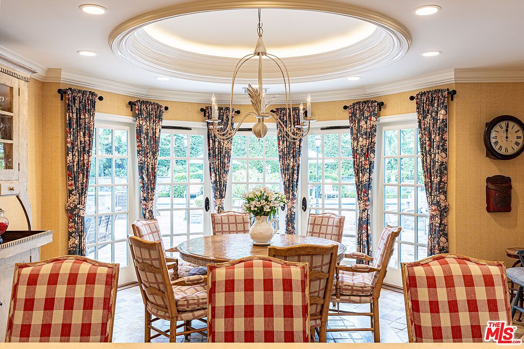 This is a dining room featuring a round wooden table surrounded by chairs with red and white checkered cushions. The room has a bright and airy feel, with large windows covered by floral curtains providing ample natural light. A chandelier hangs above the table, and the walls are painted in a warm yellow tone, creating a cozy and inviting atmosphere.
