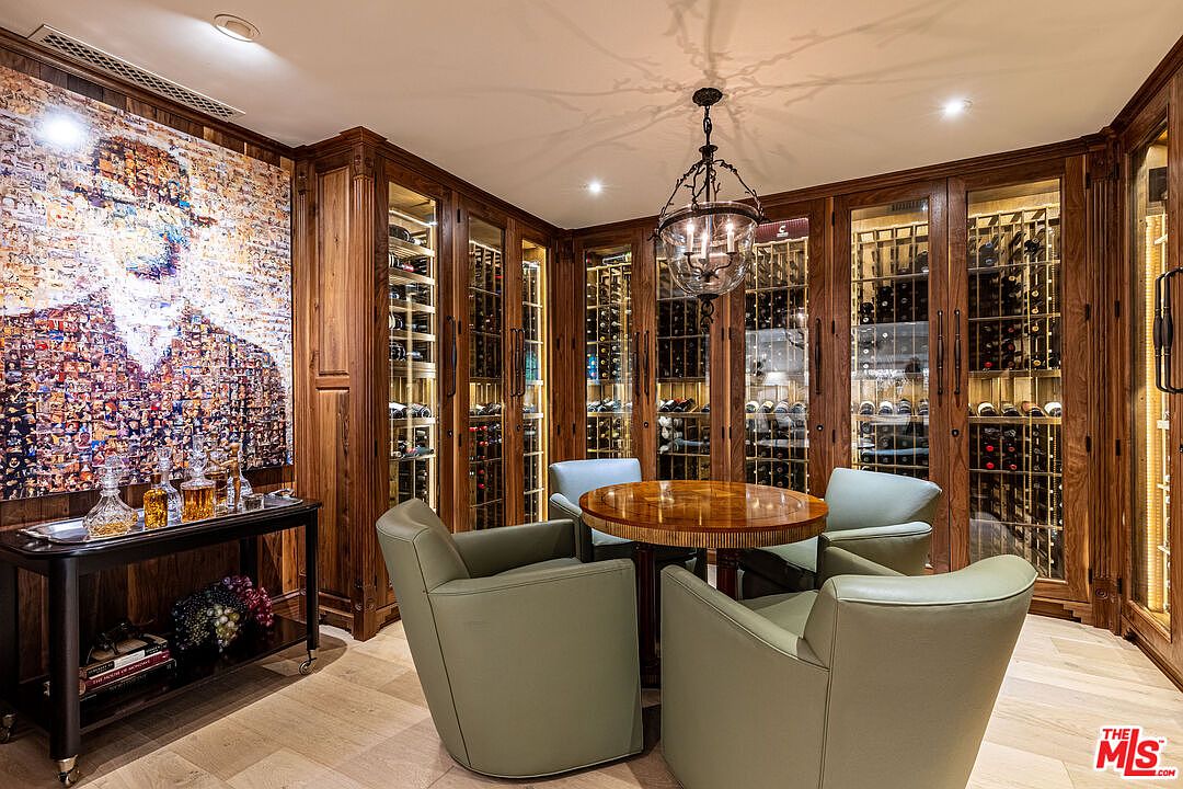 This is an elegant wine cellar featuring custom wood cabinetry with glass doors, showcasing an extensive wine collection. A round wooden table surrounded by four green leather chairs creates a cozy tasting area. The room is illuminated by a decorative chandelier and recessed lighting, enhancing the luxurious ambiance.