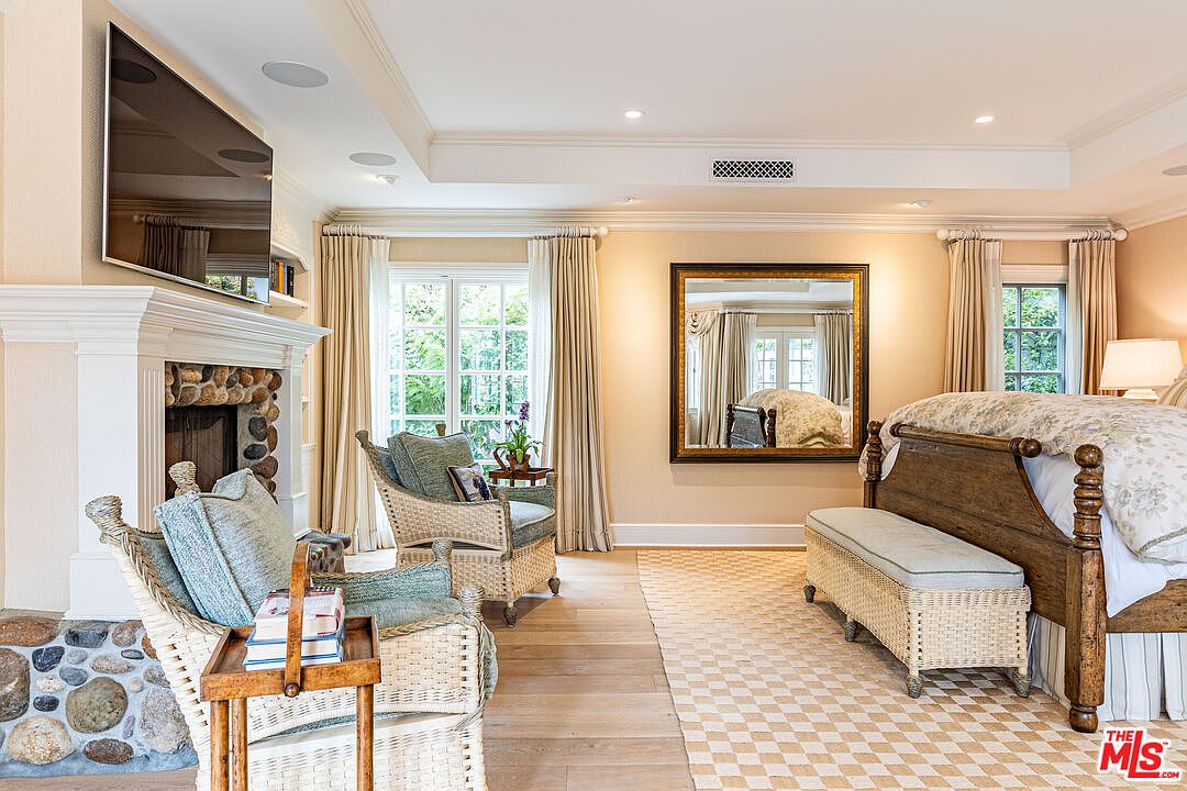 This primary bedroom features a stone fireplace with a mounted television, two wicker chairs, and a large mirror reflecting the bed. The room is decorated in neutral tones with light wood flooring and a checkered rug, creating a warm and inviting atmosphere. The perspective is from the corner of the room, showcasing the fireplace and seating area.
