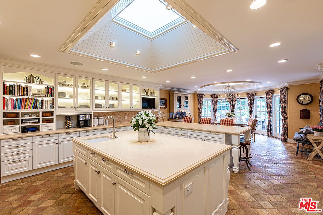 This is a bright and spacious kitchen featuring a large central island with a sink and ample counter space. The kitchen includes white cabinetry with glass-fronted display cabinets and built-in appliances. The room is well-lit with a skylight and recessed lighting, and the flooring is a patterned tile, leading to a dining area with floral curtains.