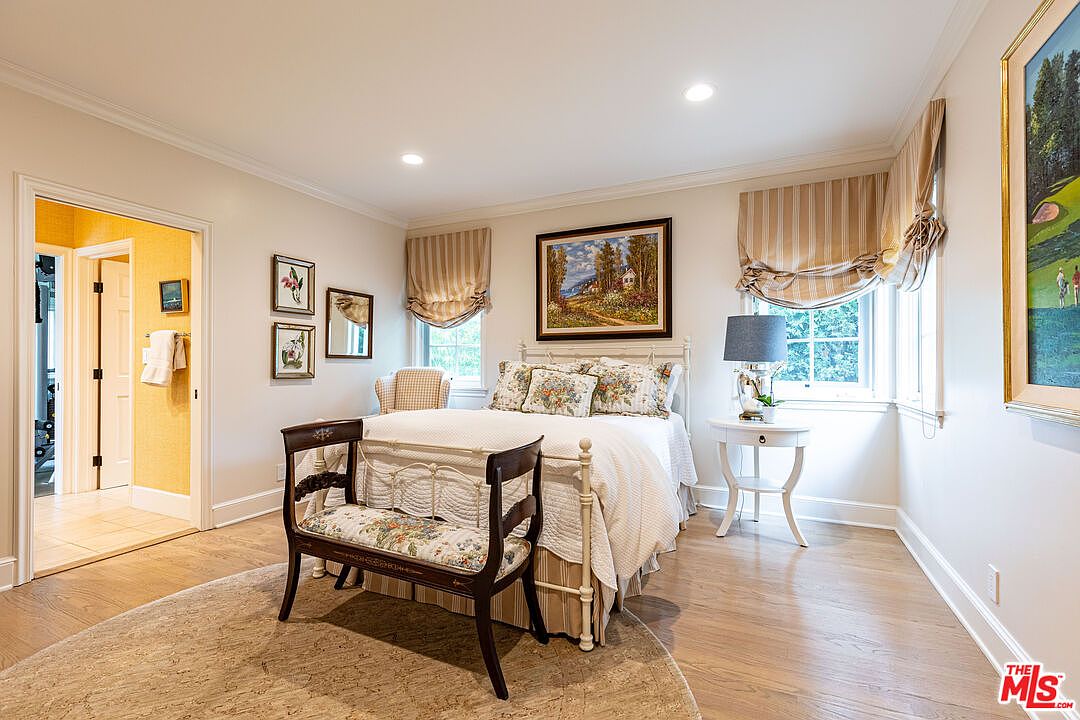 This is a cozy bedroom featuring a wrought iron bed with floral bedding, complemented by striped window treatments and framed artwork. A small bench sits at the foot of the bed, and a round rug anchors the space, creating a warm and inviting atmosphere. The room is well-lit and appears to be in excellent condition.