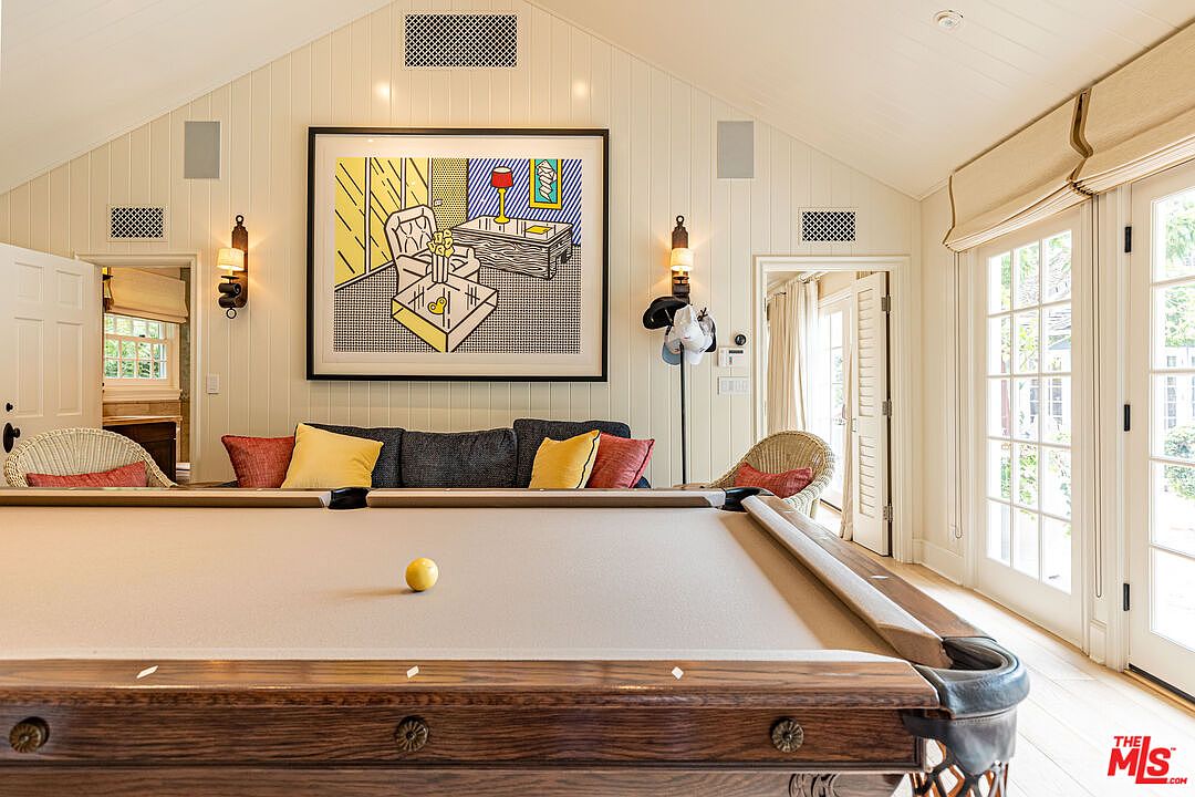 This interior shot showcases a well-lit living room featuring a pool table as a central element. The room is decorated with a large framed artwork above a sofa with colorful pillows, complemented by wall sconces and a vaulted ceiling with ventilation grilles. French doors lead to the outside, enhancing the room's bright and airy atmosphere.