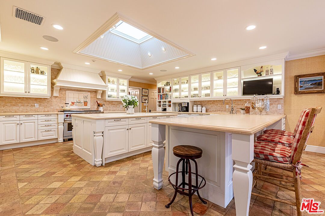 This is a spacious kitchen featuring white cabinetry with glass fronts, a large central island, and a skylight providing ample natural light. The kitchen is equipped with modern appliances and has a warm, inviting atmosphere, enhanced by the patterned tile flooring and decorative elements. A breakfast bar with seating adds to the functionality of the space.