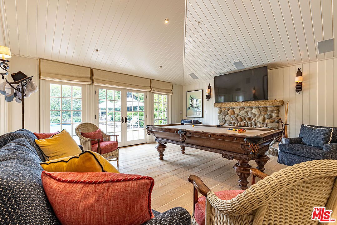 This inviting living room features a classic pool table as a centerpiece, complemented by comfortable seating arrangements including a plush sofa and wicker chairs. Natural light floods the space through large French doors, highlighting the light wood flooring and the stone-clad fireplace with a mounted television. The room exudes a relaxed and sophisticated atmosphere, perfect for entertaining or unwinding.