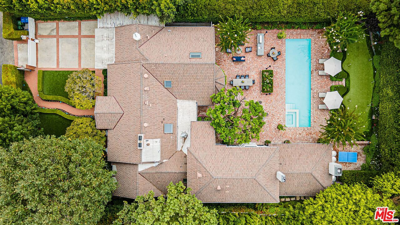 This aerial shot showcases a large, luxurious home with a sprawling layout, featuring a brown shingle roof, a rectangular swimming pool with a spa, and a brick patio area with outdoor seating. The property is surrounded by lush greenery and manicured hedges, providing privacy and a sense of seclusion. A brick driveway and walkway add to the estate's charm.