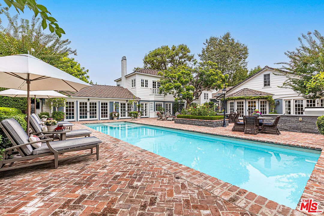 This image showcases a luxurious backyard pool area. The rectangular pool is surrounded by a brick patio, with lounge chairs and umbrellas providing a relaxing space. The house, featuring white siding and multiple windows, serves as a backdrop, complemented by lush greenery and trees.