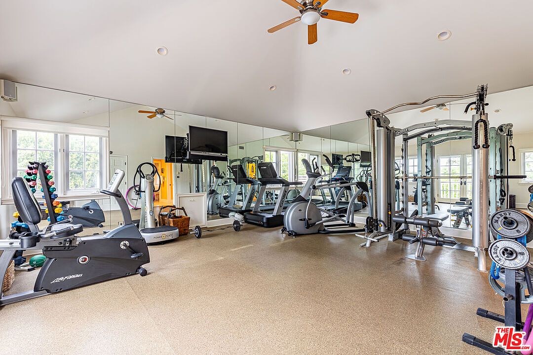 This is a spacious home gym featuring a variety of exercise equipment, including treadmills, elliptical machines, a weight rack, and a stationary bike. The room is well-lit with natural light from windows and has mirrored walls to enhance the sense of space. The flooring is a neutral color, and a ceiling fan provides ventilation.