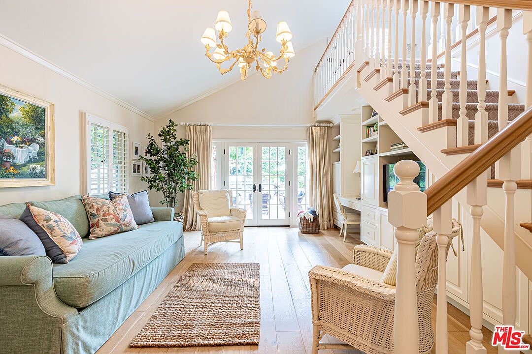 This is an interior shot of a living room featuring a light blue sofa with decorative pillows, two wicker chairs, and a jute rug on hardwood floors. The room has a high ceiling with a chandelier, and French doors lead to an outdoor area, creating a bright and airy atmosphere. A staircase with white railings and wooden handrails is visible on the right, adding architectural interest.