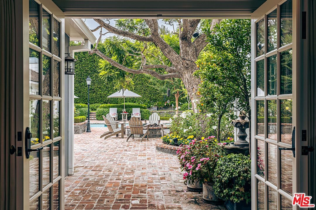 This image showcases a beautifully landscaped backyard, viewed through open French doors. The brick patio features outdoor seating around a fire pit, surrounded by lush greenery, including flowering plants and mature trees. A swimming pool is visible in the background, adding to the property's appeal as an outdoor oasis.