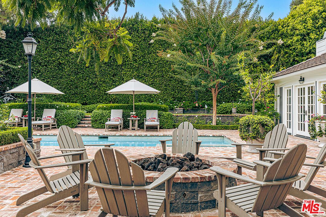 This image showcases a luxurious backyard pool and patio area. A brick fire pit surrounded by wooden Adirondack chairs takes center stage, while the pool is lined with lounge chairs and umbrellas. The space is enclosed by a tall, manicured hedge, creating a private and serene outdoor living area.