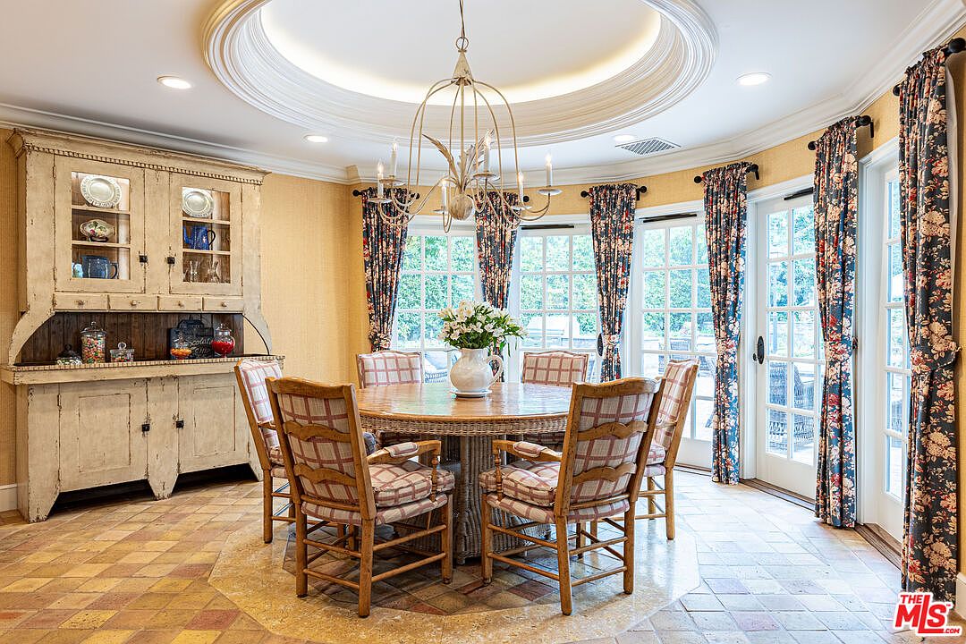This dining room features a round wicker table surrounded by six chairs with plaid cushions. A vintage hutch displays dishware, and a chandelier hangs from the decorative ceiling. Floral curtains frame the windows, adding a touch of elegance to the room.