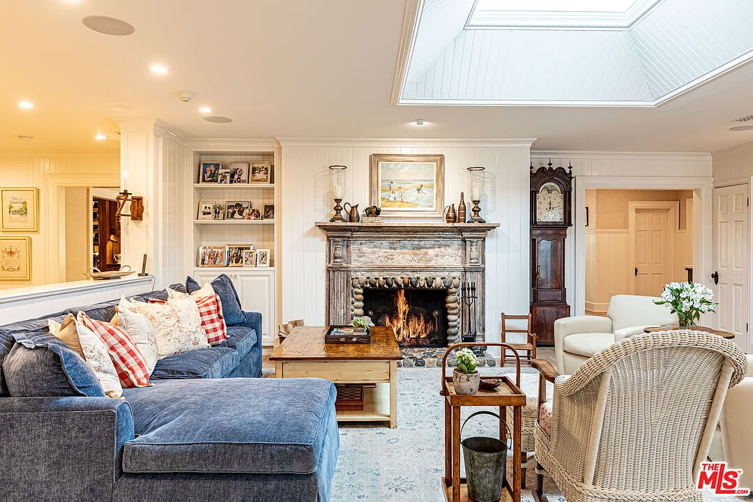 This is a cozy living room featuring a blue sectional sofa with decorative pillows, a rustic wooden coffee table, and a stone fireplace with a burning fire. The room is well-lit with natural light from a skylight and recessed lighting, and it includes built-in bookshelves and a grandfather clock, creating a warm and inviting atmosphere. The overall style is traditional with a touch of rustic charm.