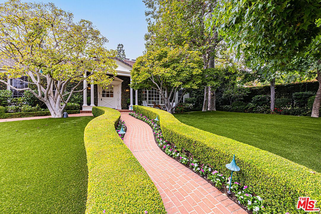 This image showcases a beautifully landscaped front yard with a winding brick pathway leading to a charming house. The pathway is bordered by meticulously manicured hedges and vibrant flower beds, creating a welcoming and picturesque scene. Lush green lawns and mature trees add to the overall appeal, suggesting a well-maintained and inviting property.