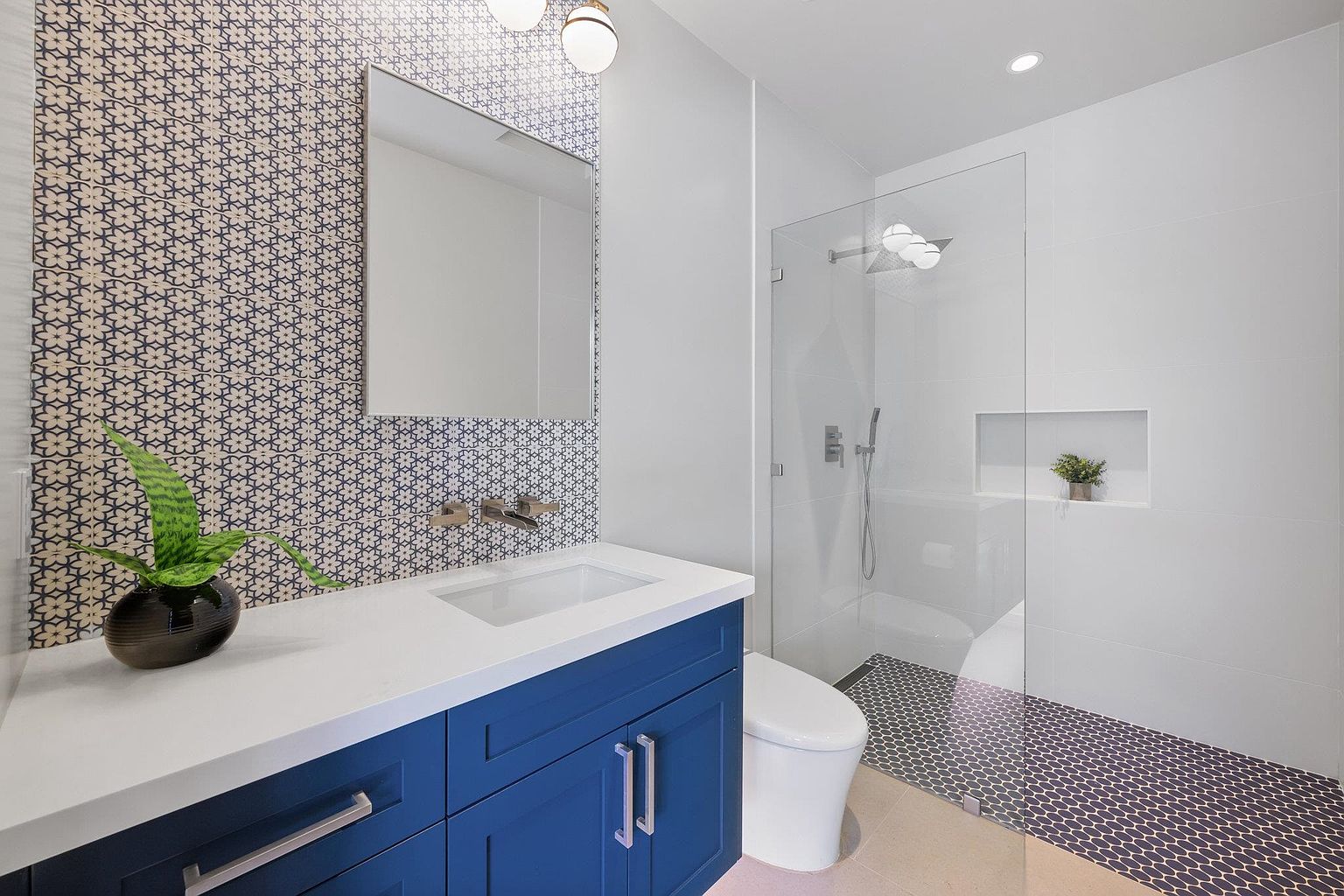 This is a modern bathroom featuring a blue vanity with a white countertop and a rectangular sink. The wall behind the vanity is decorated with patterned tiles, and a mirror hangs above the sink. A glass-enclosed shower is visible to the right, with a built-in niche for toiletries. The floor is a combination of light-colored tiles and patterned tiles inside the shower area.