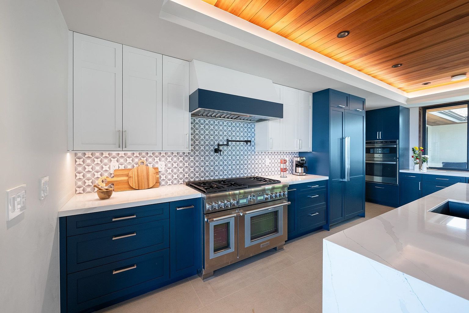 This is a modern kitchen with two-tone cabinetry, featuring white upper cabinets and navy blue lower cabinets. Stainless steel appliances, including a large range and refrigerator, add a sleek touch. The backsplash is a patterned tile, and the countertops are a light-colored stone, with a wood ceiling adding warmth to the space. The perspective is from a medium angle, showcasing the layout and design elements.