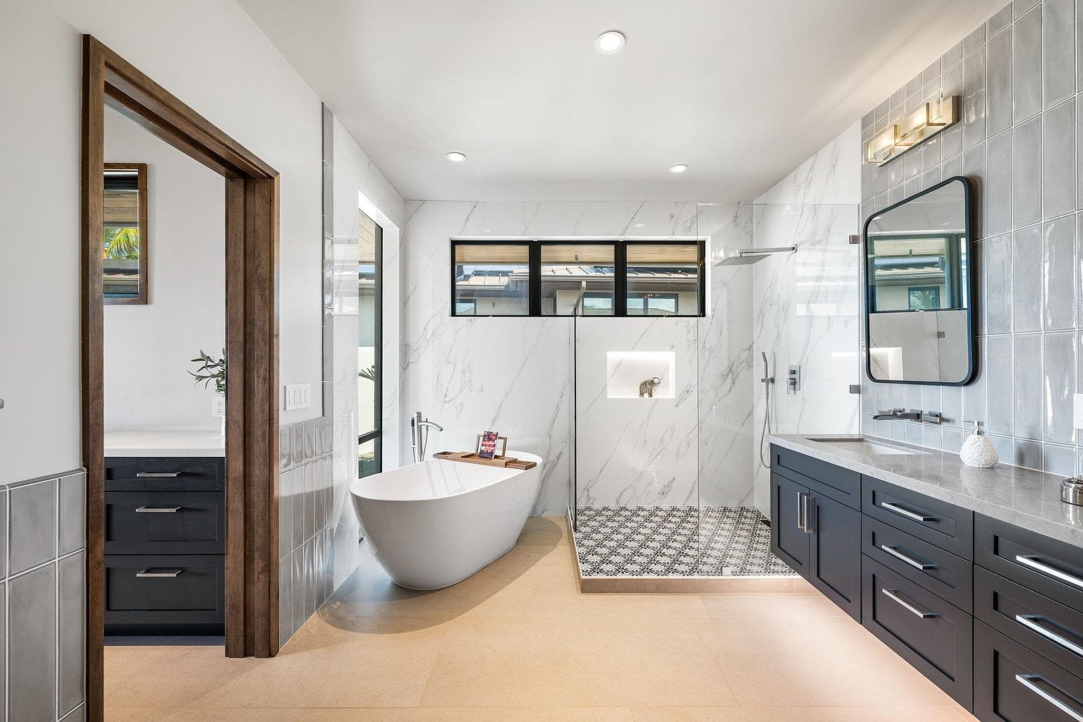 This is a modern primary bathroom featuring a freestanding bathtub, a walk-in shower with marble tile, and a dark vanity with a light countertop. The bathroom has a neutral color palette with gray and white tones, creating a spa-like atmosphere. The space is well-lit with recessed lighting and a window above the tub.