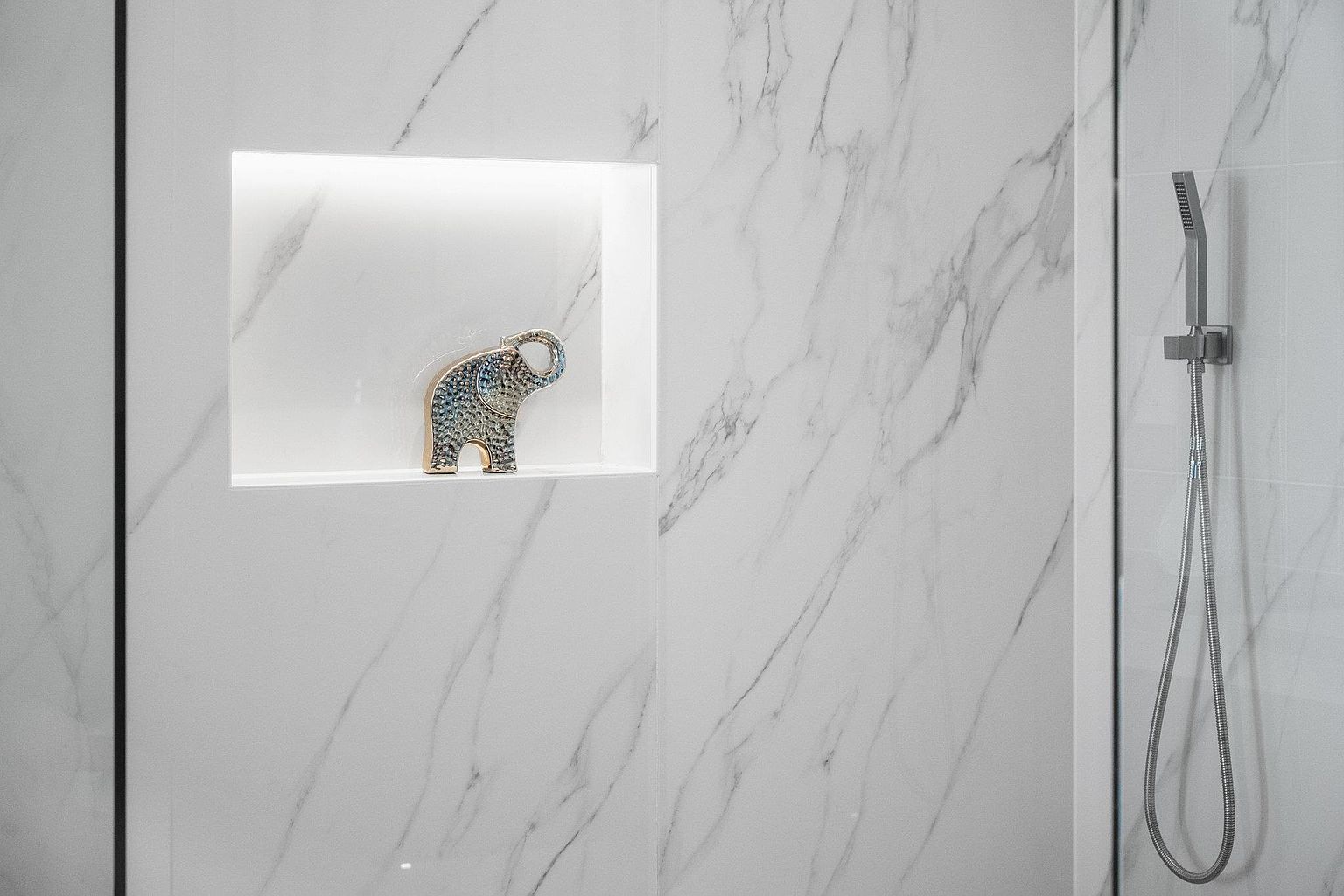 This image showcases a modern primary bathroom featuring white marble walls with subtle gray veining. A built-in niche houses a decorative elephant figurine, illuminated by soft lighting. A sleek, minimalist showerhead and hose are visible on the right, contributing to the bathroom's clean and luxurious aesthetic.