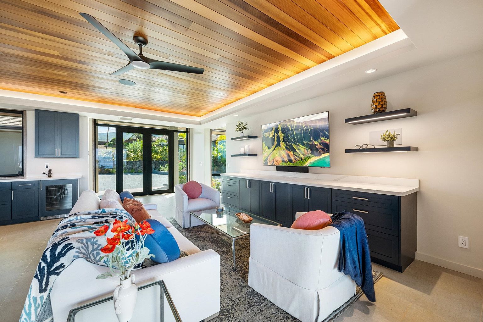 This living room features a modern design with a wood-paneled ceiling and recessed lighting. The room includes a built-in bar area, a large television, and comfortable white seating arranged around a glass coffee table. Large glass doors provide access to an outdoor area, enhancing the indoor-outdoor flow.