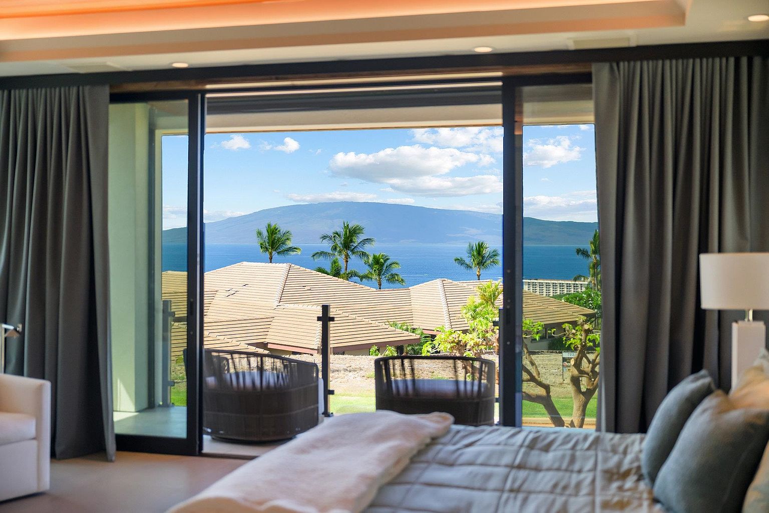This is a view from a primary bedroom, showcasing a sliding glass door that opens to a balcony with seating. The room features a large bed with plush bedding and neutral tones. The view outside includes rooftops, palm trees, the ocean, and a distant island, creating a serene and luxurious atmosphere.