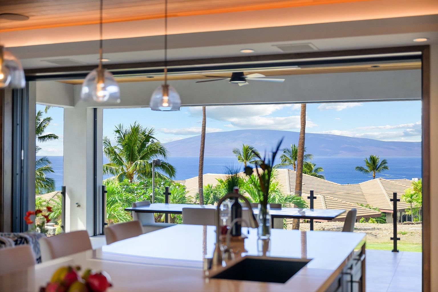 This image showcases a luxurious kitchen with a stunning view of the ocean and a distant island. The kitchen features a large island with a sink and modern fixtures, complemented by pendant lighting. The open-concept design seamlessly blends indoor and outdoor living, creating a serene and upscale atmosphere.
