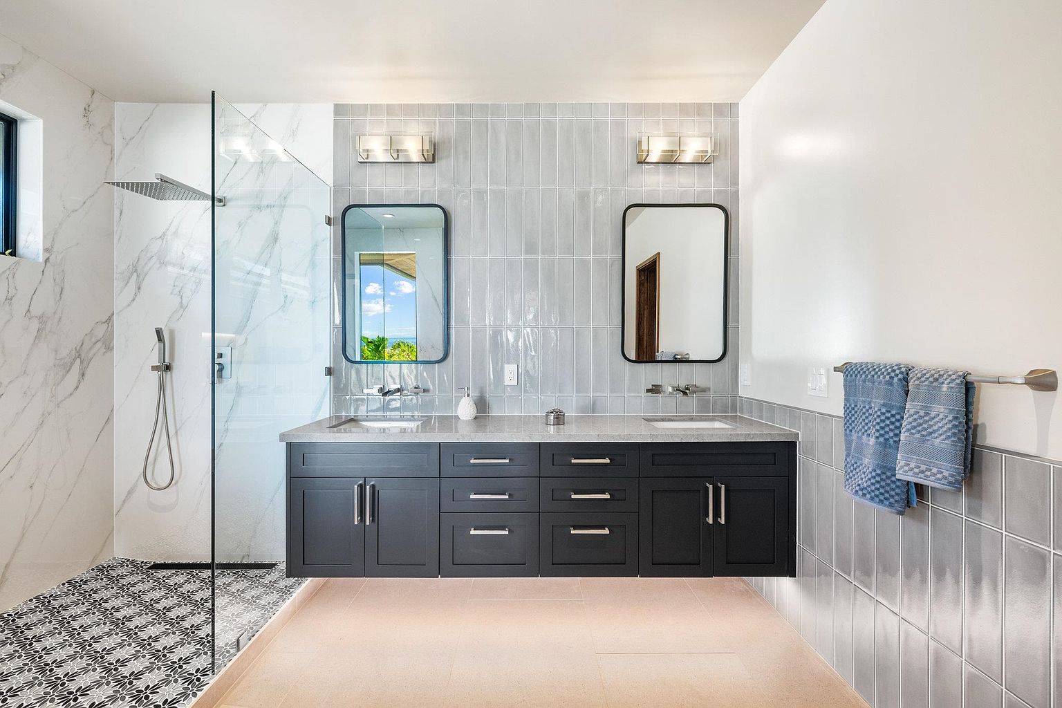 This is a well-lit primary bathroom featuring a double vanity with dark cabinetry and light countertops. Two rectangular mirrors hang above the sinks, illuminated by modern sconces. A glass-enclosed shower with marble-like walls and patterned floor tiles adds a luxurious touch, while the overall design is clean and contemporary.