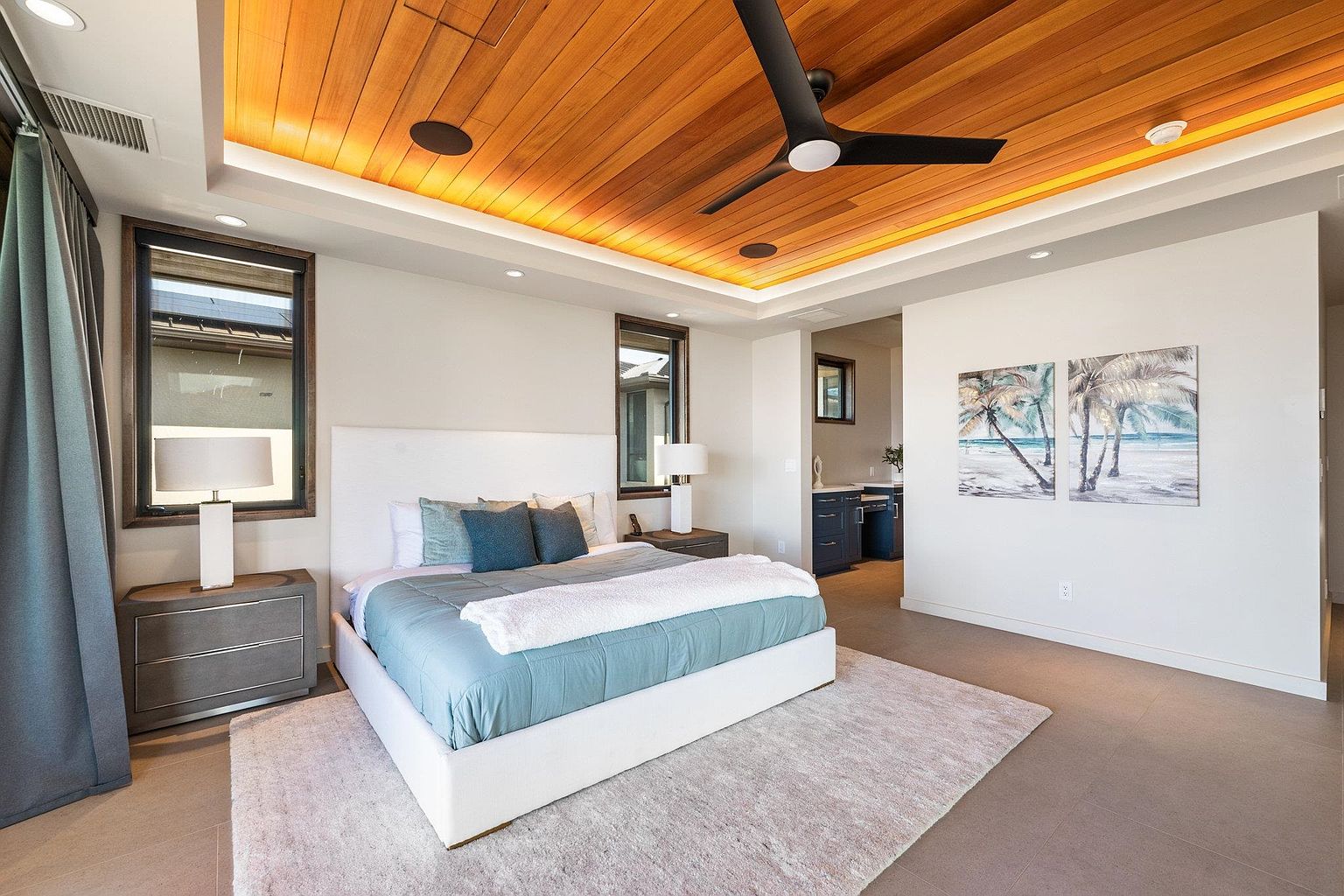 This is a primary bedroom featuring a king-size bed with a white upholstered headboard, blue bedding, and a white throw. The room has a wood-paneled ceiling with recessed lighting and a modern ceiling fan. There are two windows, one on each side of the bed, and two pieces of artwork on the wall. The perspective is from the foot of the bed, looking towards the headboard and the rest of the room.