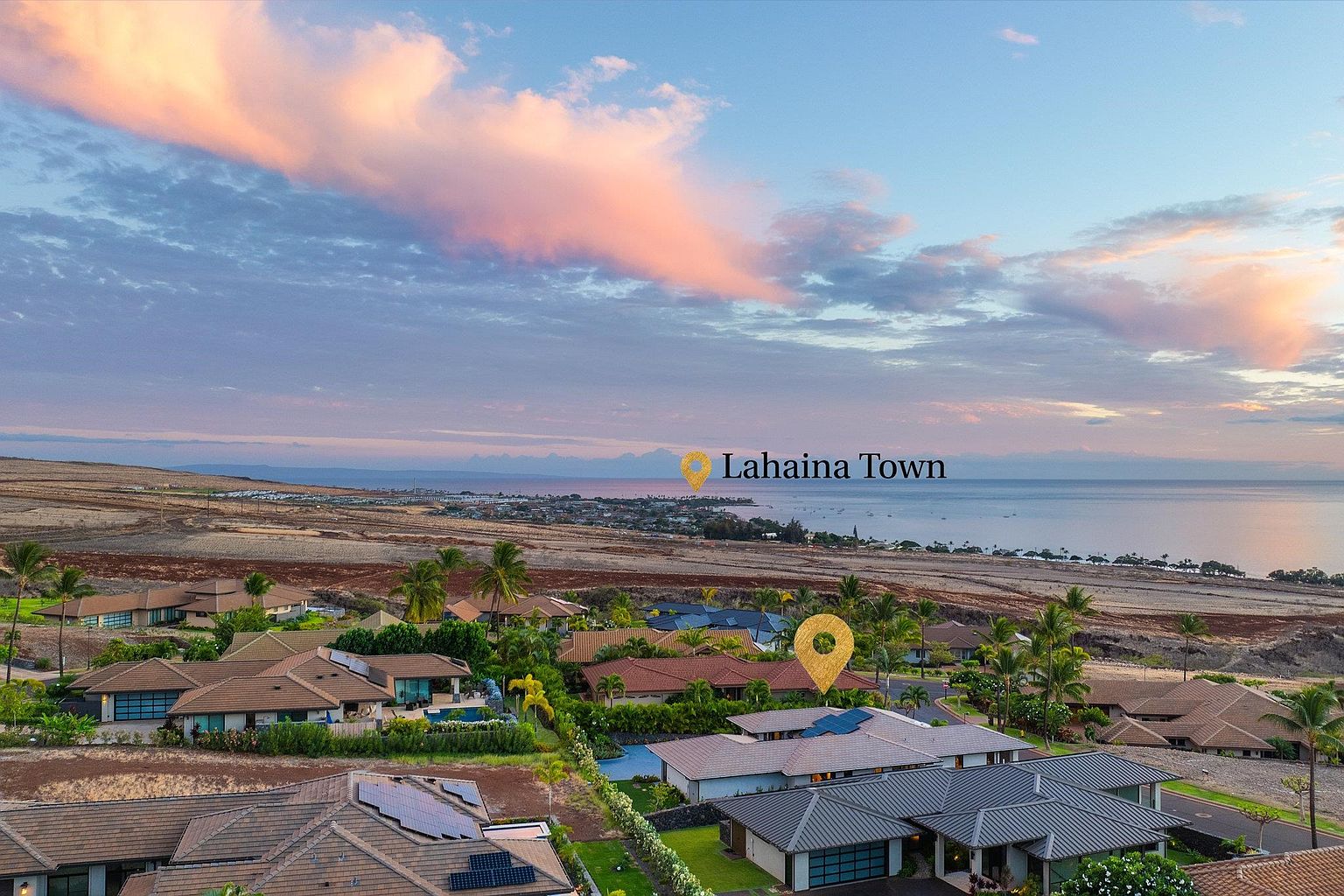 This aerial view showcases a residential neighborhood with well-maintained homes, lush landscaping, and a glimpse of the ocean in the background. The houses feature varied rooflines and architectural styles, some with solar panels, suggesting a modern and eco-conscious community. The overall impression is one of a desirable and upscale living environment.