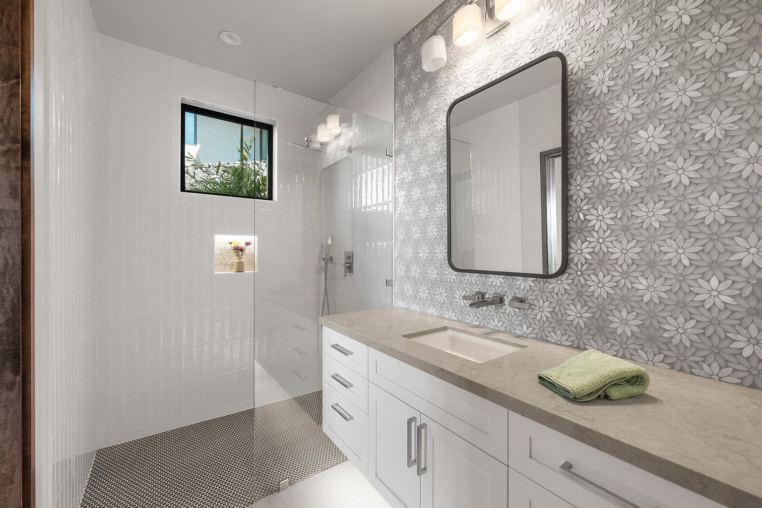 This is a well-lit bathroom featuring a modern design. The space includes a vanity with white cabinets and a light-colored countertop, a rectangular mirror with a black frame, and a shower area enclosed by glass. The wall behind the vanity is adorned with a floral-patterned tile, while the shower area has white tiles and a small window.