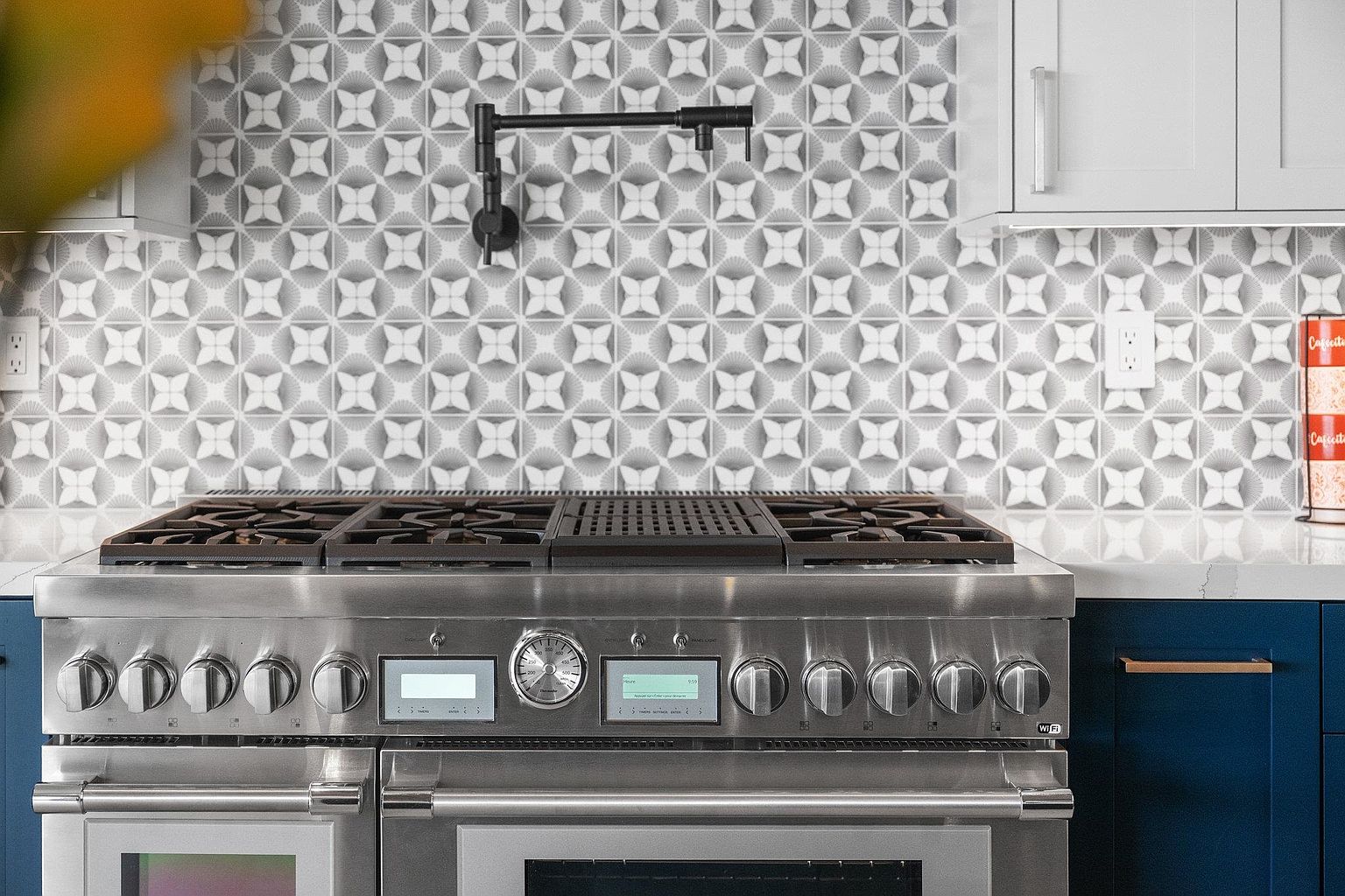 This is a close-up shot of a modern kitchen featuring a stainless steel range with multiple burners and digital displays. The backsplash is a patterned gray and white tile, complemented by a black pot filler faucet. The cabinetry is a mix of white and blue, with a white countertop.