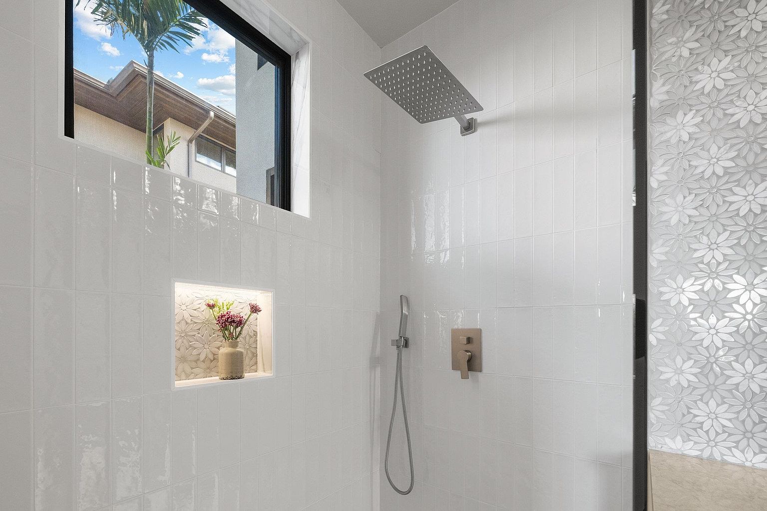 This is a bright and modern primary bathroom featuring white tiled walls, a rainfall showerhead, and a handheld shower fixture. A niche in the wall displays decorative flowers, adding a touch of elegance. A window provides natural light and a view to the outside, enhancing the spa-like atmosphere.