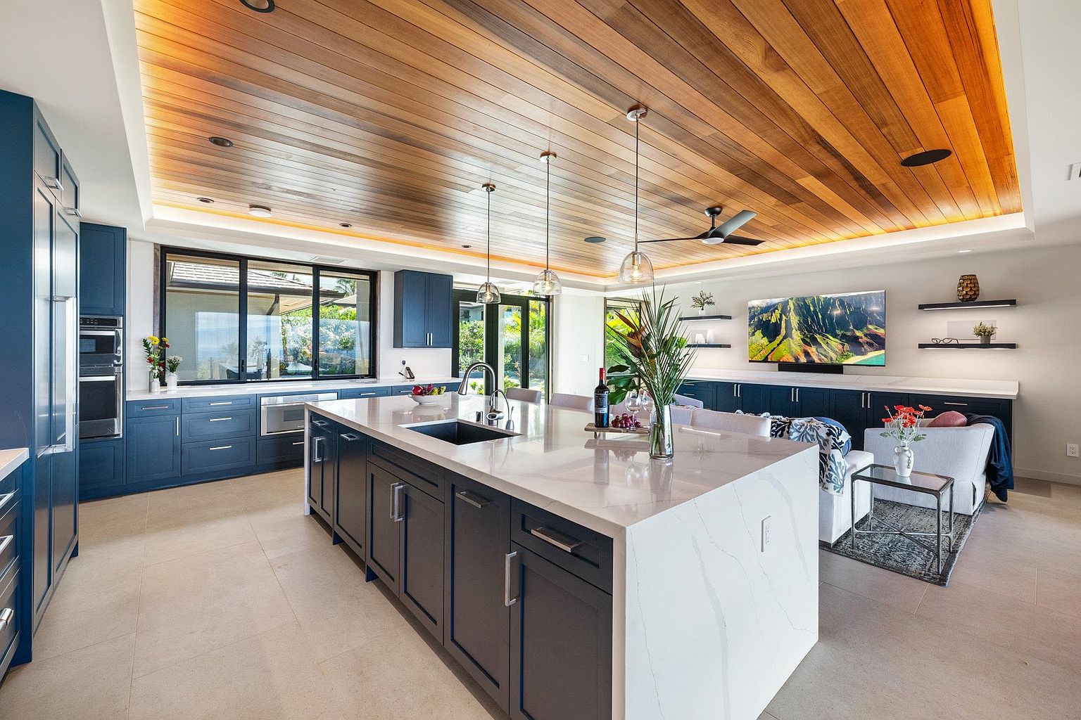 This is a spacious, modern kitchen featuring dark blue cabinetry, a large white quartz island with a sink, and stainless steel appliances. The ceiling is wood paneled with recessed lighting, and pendant lights hang above the island. The kitchen seamlessly transitions into a living area with a sofa and television, creating an open-concept living space.