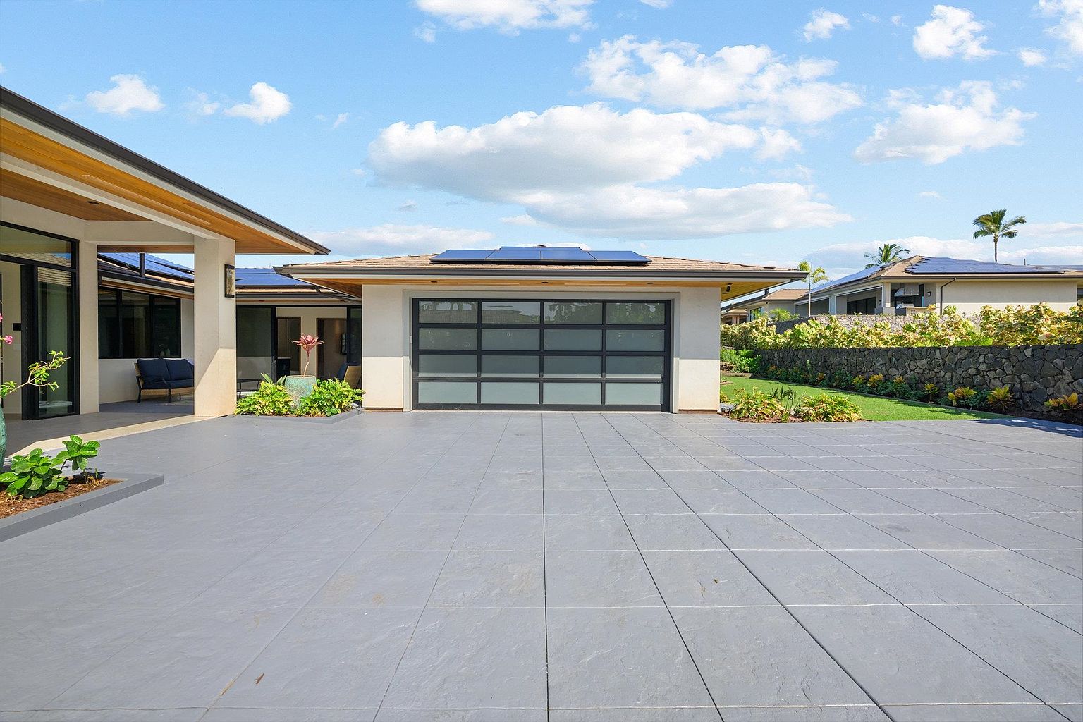 The image showcases a modern garage with a sleek, frosted glass door framed by dark trim, set against a light-colored exterior. Solar panels are visible on the roof, indicating energy efficiency. The driveway is paved with large gray tiles, and landscaping adds a touch of greenery to the scene.