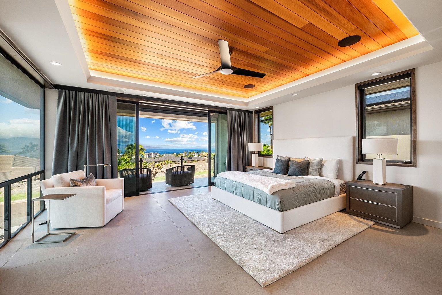 This is a luxurious primary bedroom featuring a king-size bed with a white upholstered headboard, a gray and white color scheme, and a stunning ocean view through sliding glass doors. The room includes a seating area with a white armchair, a wooden ceiling with recessed lighting, and a modern nightstand with a lamp. The overall impression is one of elegance and tranquility.