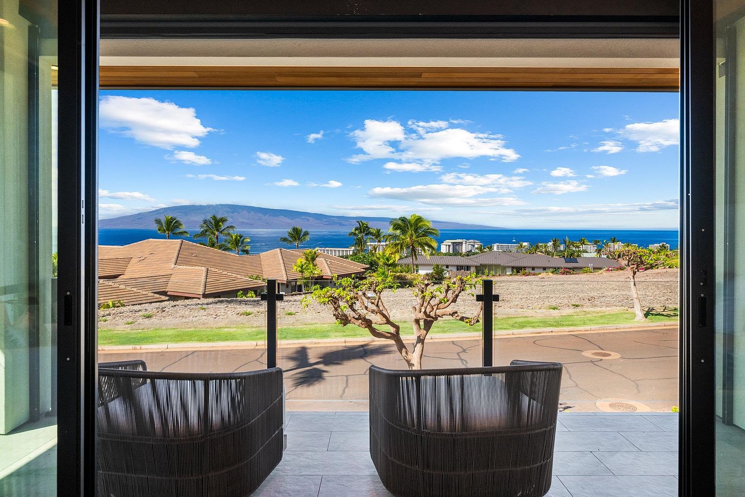 This image showcases a luxurious balcony with two modern chairs, offering a stunning panoramic view of the ocean, a distant island, and nearby residential rooftops. The open sliding doors create a seamless transition between indoor and outdoor living, emphasizing the property's appeal for relaxation and enjoying the scenic surroundings. The balcony's tile flooring adds a touch of elegance.